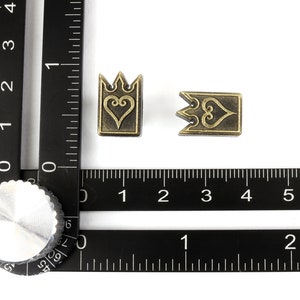 May include: Two antique brass-colored metal charms. One charm is a crown shape with a heart design, and the other is a rectangular shape with a heart design. The charms are approximately 0.5 inches in size.