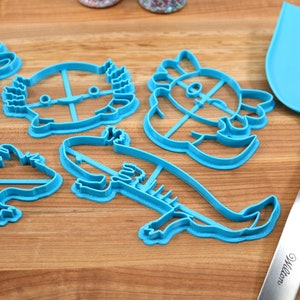 May include: Five blue plastic cookie cutters shaped like different animals, including a turtle, a dragon, a fish, and two axolotls. A blue spatula with a silver handle is also in the image.