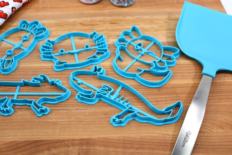 May include: Five blue plastic cookie cutters shaped like different animals, including a turtle, a dragon, a fish, and two axolotls. A blue spatula with a silver handle is also in the image.