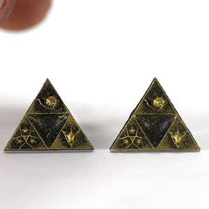 May include: A pair of gold-colored triangular earrings with a black background and intricate designs. The earrings feature a triangular pattern with a star, a crescent moon, and a stylized bird.