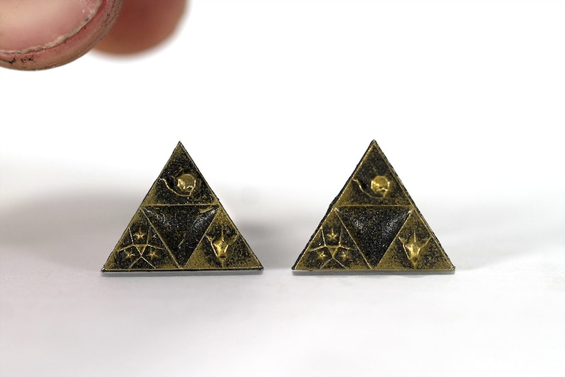 May include: A pair of gold-colored triangular earrings with a black background and intricate designs. The earrings feature a triangular pattern with a star, a crescent moon, and a stylized bird.