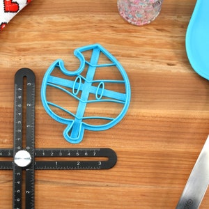 May include: A blue plastic cookie cutter shaped like a leaf with a face. The cutter is on a wooden surface with a ruler and a blue spatula.