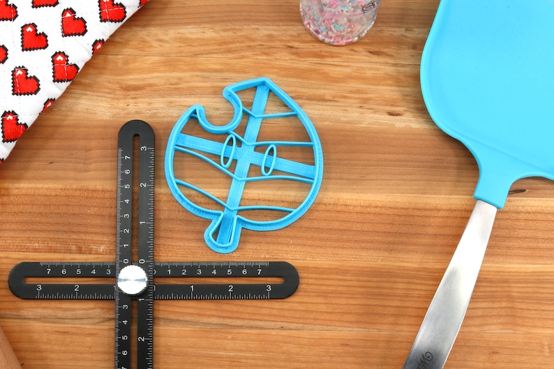May include: A blue plastic cookie cutter shaped like a leaf with a face. The cutter is on a wooden surface with a ruler and a blue spatula.