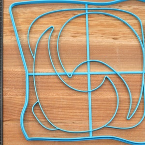May include: A blue plastic mold for making a decorative design in food. The mold is shaped like a square with a curved design inside.