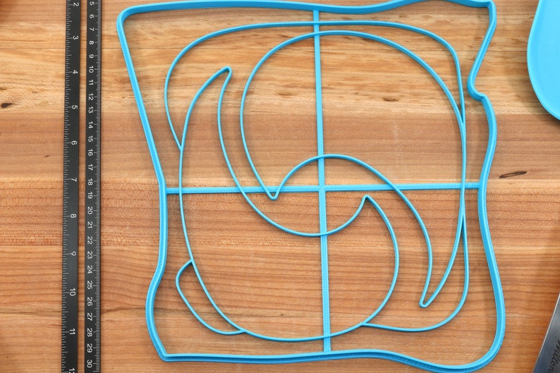 May include: A blue plastic mold for making a decorative design in food. The mold is shaped like a square with a curved design inside.