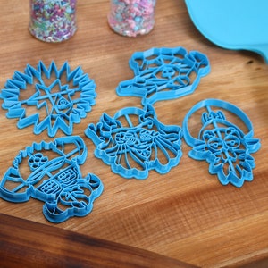 May include: Six blue plastic cookie cutters on a wooden surface. The cutters are shaped like a star, a face, a flower, and two other abstract designs.
