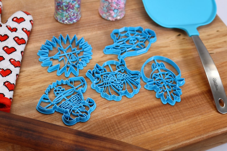 May include: Six blue plastic cookie cutters on a wooden surface. The cutters are shaped like a star, a face, a flower, and two other abstract designs.