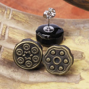 以下が含まれることがあります： アンティークスタイルのスタッドピアス。銃のシリンダーのデザイン。ピアスはブロンズ色で、詳細な模様があります。片方のピアスは、銀色のポストが付いた黒いスタッドに取り付けられています。木製の表面に展示されています。