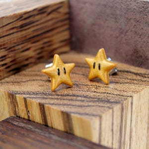 May include: A pair of gold star-shaped earrings with black accents. The earrings are on a wooden surface.