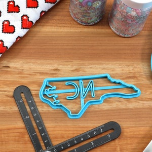 May include: Blue plastic cookie cutter shaped like the state of North Carolina with the letters "NC" inside.