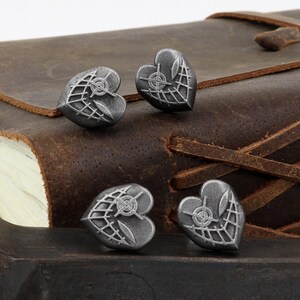 May include: Four silver heart-shaped charms with a detailed design of a crosshair and a web pattern. The charms are on a brown leather surface.
