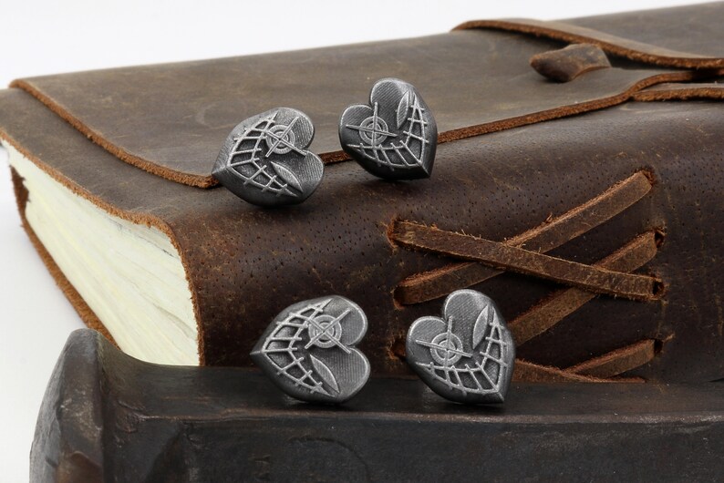 May include: Four silver heart-shaped charms with a detailed design of a crosshair and a web pattern. The charms are on a brown leather surface.