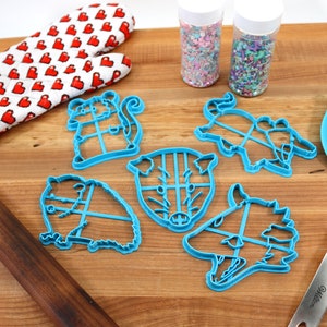 May include: A set of six blue plastic animal cookie cutters on a wooden surface. The cutters are shaped like a pig, a wolf, a bear, a squirrel, a deer, and a rhinoceros. There are also two jars of sprinkles, a rolling pin, a spatula, and a red oven mitt with white hearts.
