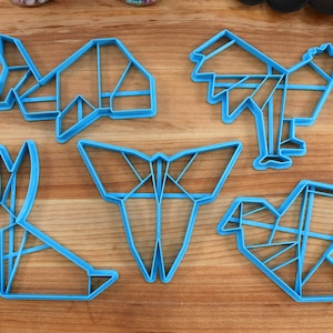 May include: Five blue plastic cookie cutters shaped like origami animals: a fish, a rooster, a butterfly, a rabbit, and a bird.