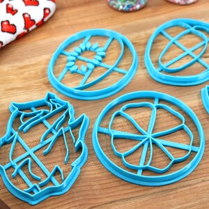 May include: Five blue plastic cookie cutters in different shapes, including a flower, a sun, a leaf, and two abstract shapes. The cutters are on a wooden surface.