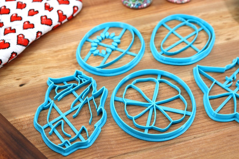 May include: Five blue plastic cookie cutters in different shapes, including a flower, a sun, a leaf, and two abstract shapes. The cutters are on a wooden surface.