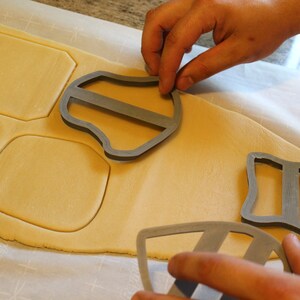 May include: A person is using cookie cutters to cut out shapes from a sheet of cookie dough. The cookie cutters are made of metal and have different shapes, including a shield, a square, and a rectangle. The cookie dough is a light brown color and is on a white surface.