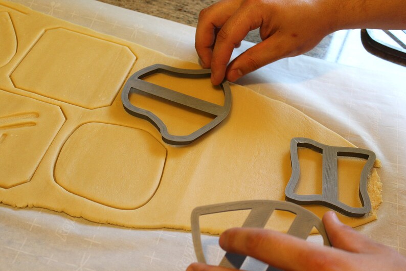 May include: A person is using cookie cutters to cut out shapes from a sheet of cookie dough. The cookie cutters are made of metal and have different shapes, including a shield, a square, and a rectangle. The cookie dough is a light brown color and is on a white surface.