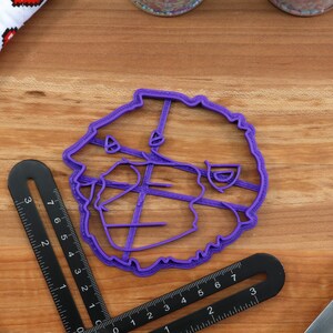 May include: Purple plastic cookie cutter shaped like a cartoon bear's head. The cutter has a detailed design with lines and curves.