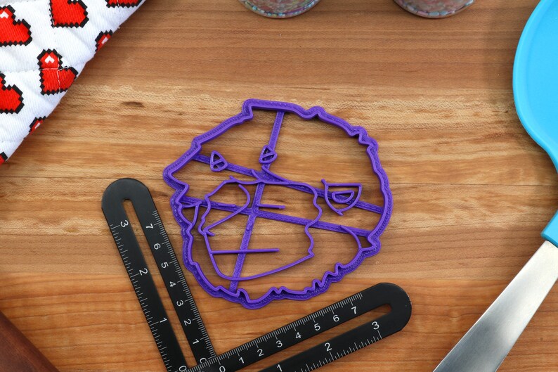 May include: Purple plastic cookie cutter shaped like a cartoon bear's head. The cutter has a detailed design with lines and curves.