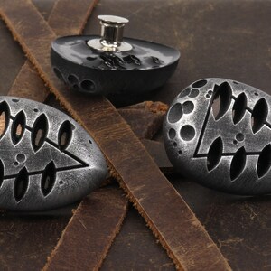 May include: Three silver-toned metal buttons with a geometric design. The buttons are shaped like a teardrop and have a raised, textured surface. The design features a triangle with a series of oval cutouts.