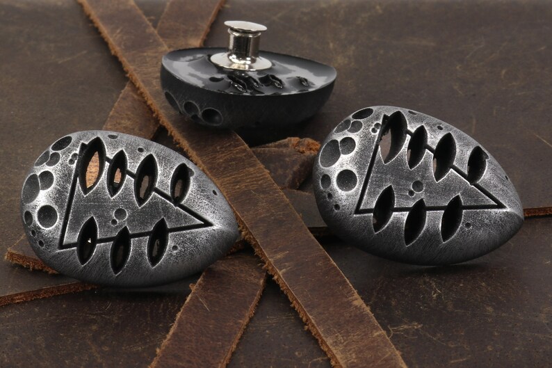 May include: Three silver-toned metal buttons with a geometric design. The buttons are shaped like a teardrop and have a raised, textured surface. The design features a triangle with a series of oval cutouts.