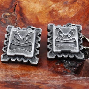 May include: Two silver-toned metal keychains with a square shape and a detailed design of a frowning face. The keychains are on a wooden surface.