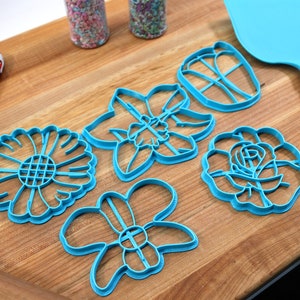 May include: Set of five blue plastic flower-shaped cookie cutters on a wooden surface. The cutters are shaped like a daisy, a daffodil, a butterfly, a rose, and a generic flower.
