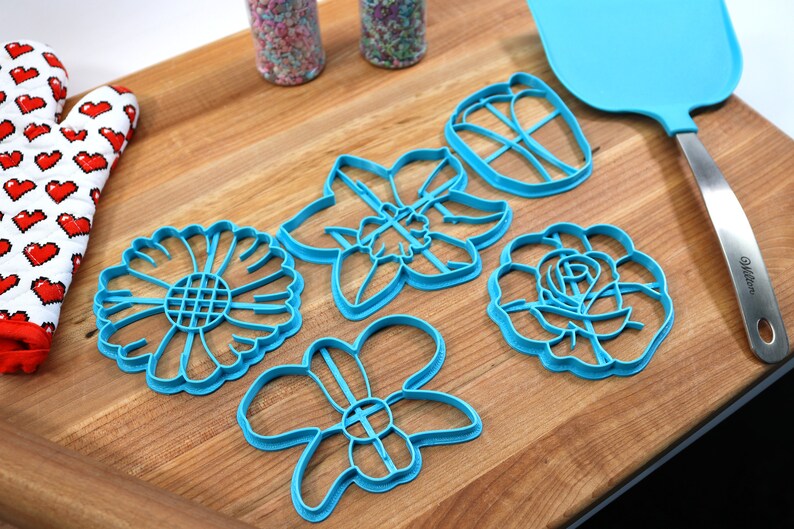May include: Set of five blue plastic flower-shaped cookie cutters on a wooden surface. The cutters are shaped like a daisy, a daffodil, a butterfly, a rose, and a generic flower.