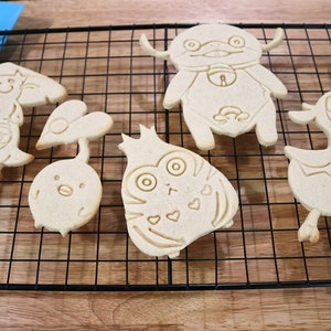 May include: Six different cookie cutters on a black wire rack. The cookie cutters are shaped like various cartoon characters, including a duck, a bird, a cat, and a bear.
