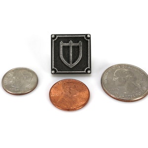 Puede incluir: Un pin de metal plateado con un fondo cuadrado negro y un dise&ntilde;o de escudo plateado. El pin se muestra junto a tres monedas estadounidenses: una moneda de veinticinco centavos, un centavo y una moneda de diez centavos.