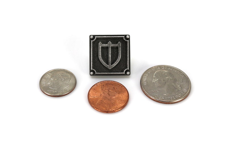 Puede incluir: Un pin de metal plateado con un fondo cuadrado negro y un dise&ntilde;o de escudo plateado. El pin se muestra junto a tres monedas estadounidenses: una moneda de veinticinco centavos, un centavo y una moneda de diez centavos.