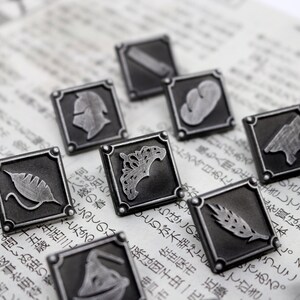 May include: A set of ten silver-toned metal pins with different designs. Each pin features a square shape with a raised design inside. The designs include a crown, a leaf, a wheat stalk, a helmet, and other abstract shapes.