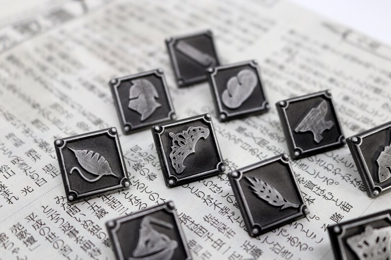 May include: A set of ten silver-toned metal pins with different designs. Each pin features a square shape with a raised design inside. The designs include a crown, a leaf, a wheat stalk, a helmet, and other abstract shapes.
