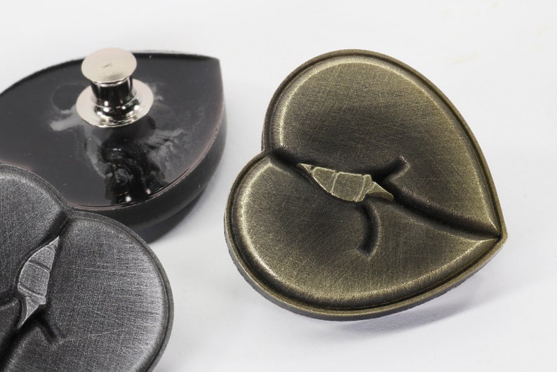 May include: Three heart-shaped metal buttons. One button is black with a silver stud, one is black with a crack, and one is gold with a crack.