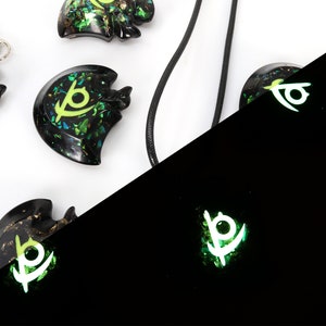 May include: A set of four black resin pendants with a green and gold glitter design. The pendants feature a stylized eye symbol. The pendants are shown in both regular and glow-in-the-dark lighting.