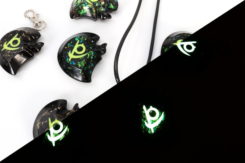May include: A set of four black resin pendants with a green and gold glitter design. The pendants feature a stylized eye symbol. The pendants are shown in both regular and glow-in-the-dark lighting.