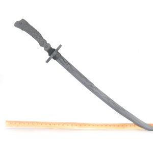 Może przedstawiać: Szary miecz katana z teksturowanym uchwytem i wzorem ostrza. Miecz ma około 76 cm długości. Drewniana linijka jest umieszczona obok miecza dla skali, na białym tle.