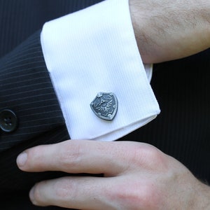 May include: A pair of silver cufflinks featuring a stylised shield with a winged eye design.