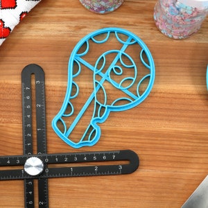 May include: A blue plastic cookie cutter shaped like a candy cane with a swirl design. The cutter is on a wooden surface with a ruler and a blue spatula.