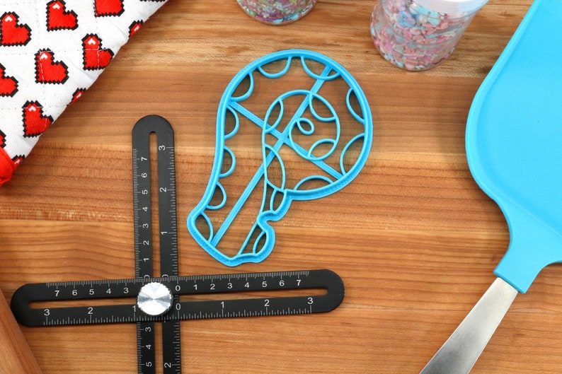 May include: A blue plastic cookie cutter shaped like a candy cane with a swirl design. The cutter is on a wooden surface with a ruler and a blue spatula.