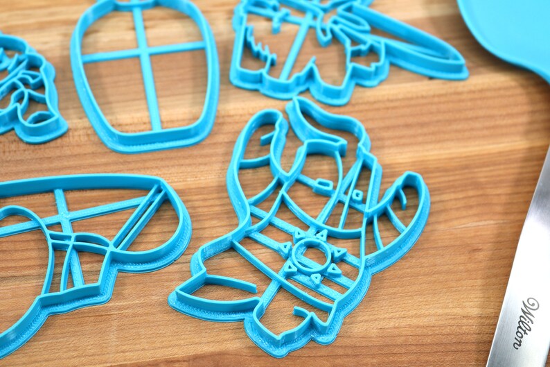 May include: A set of blue plastic cookie cutters shaped like a llama, a cactus, and a bottle. The cutters are on a wooden surface.