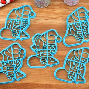 May include: Five blue plastic cookie cutters shaped like cartoon characters wearing Santa hats and scarves. The characters are holding a snowball.