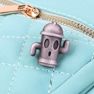 May include: A silver-coloured metal pin shaped like a fire hydrant with a cartoon face. The fire hydrant has two arms and a surprised expression.