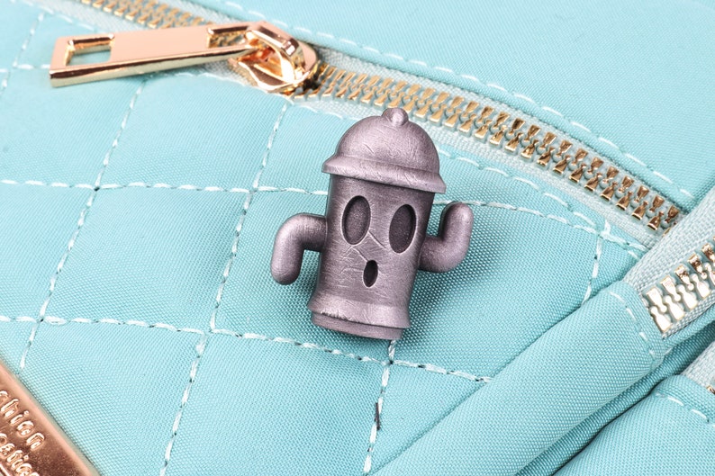 May include: A silver-coloured metal pin shaped like a fire hydrant with a cartoon face. The fire hydrant has two arms and a surprised expression.