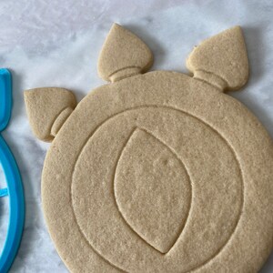 May include: A plain, light brown sugar cookie in the shape of a circular shield with three points at the top. The cookie has a crescent moon design in the center.