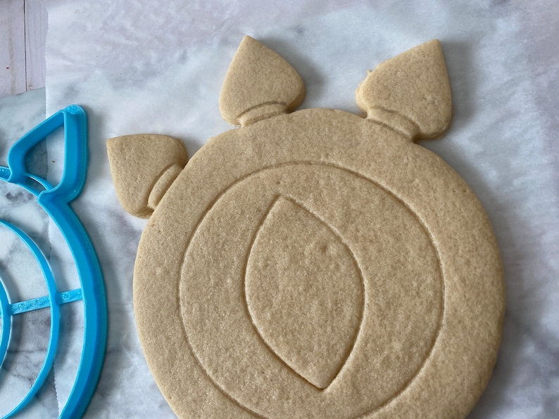 May include: A plain, light brown sugar cookie in the shape of a circular shield with three points at the top. The cookie has a crescent moon design in the center.