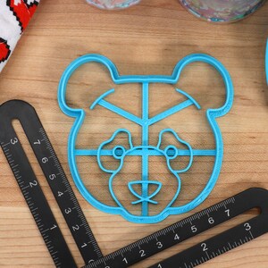 May include: A blue plastic cookie cutter shaped like a bear's face. The cutter has a grid pattern inside the bear's face.