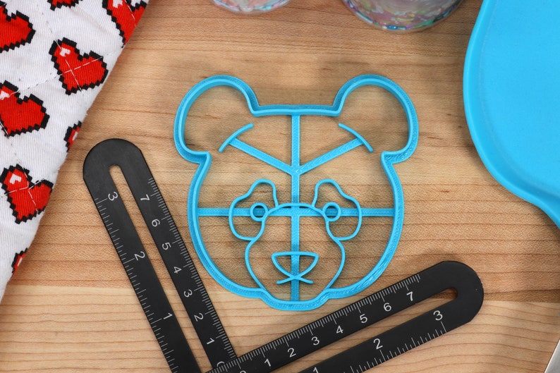 May include: A blue plastic cookie cutter shaped like a bear's face. The cutter has a grid pattern inside the bear's face.