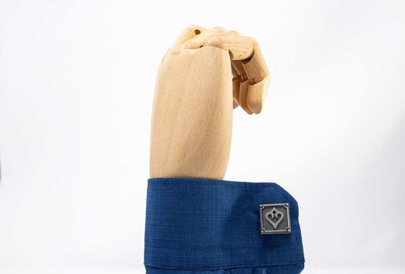 May include: A pair of silver cufflinks with a heart design. The cufflinks are on a blue shirt cuff.